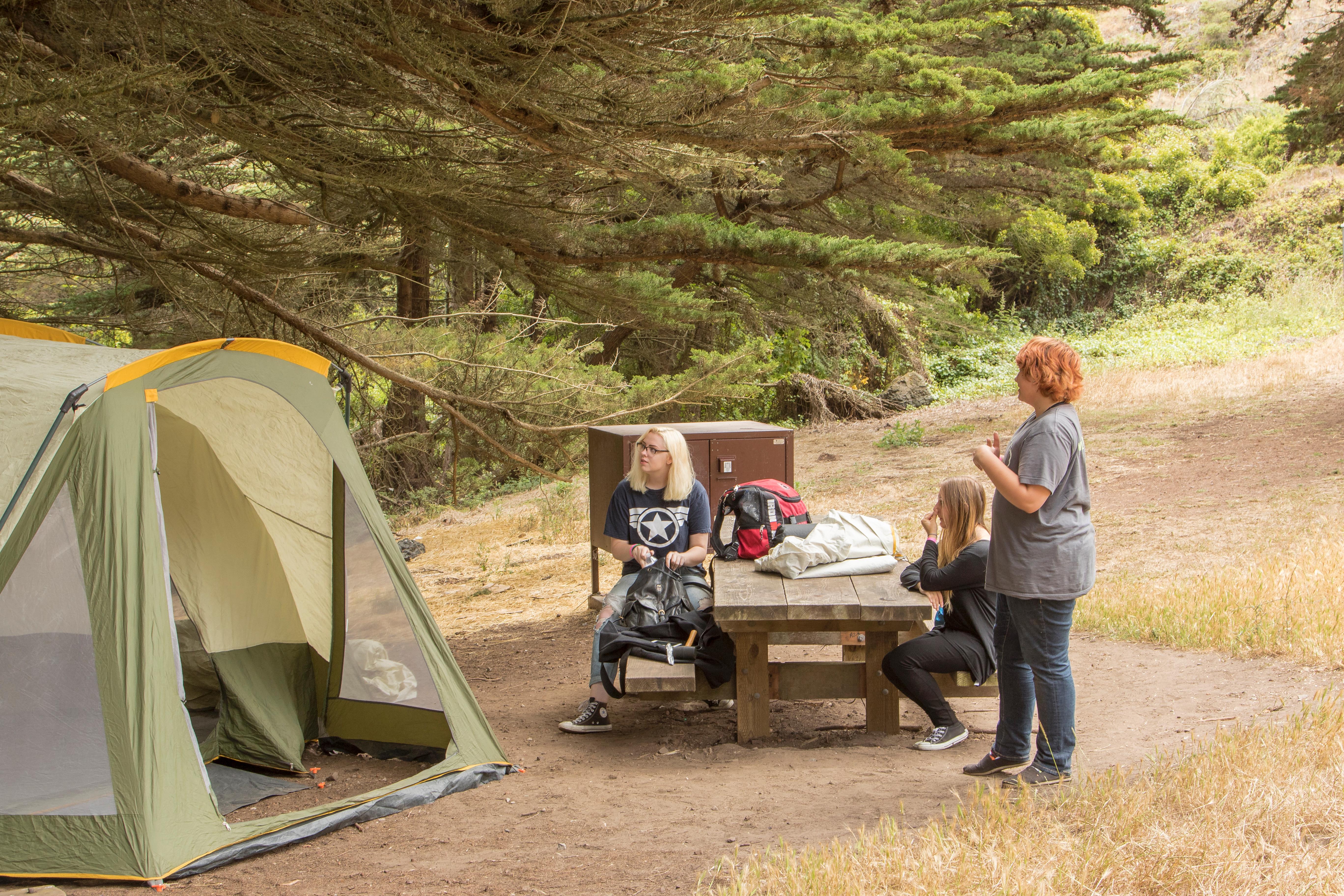 Campers have their tent set up next to a picnic table covered in food and supplies.