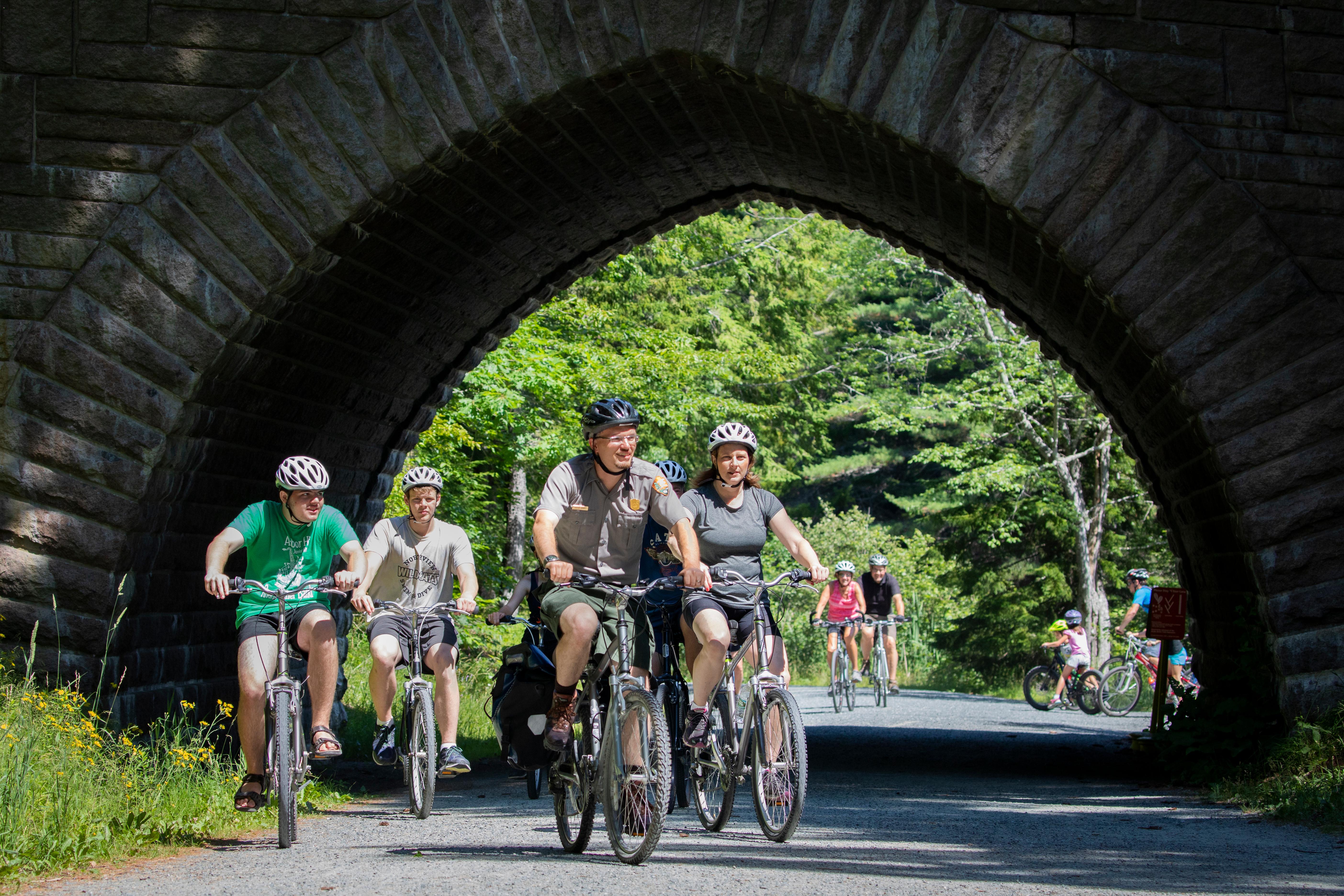 A group of people riding bicycles travels under the arch of a large stone bridge