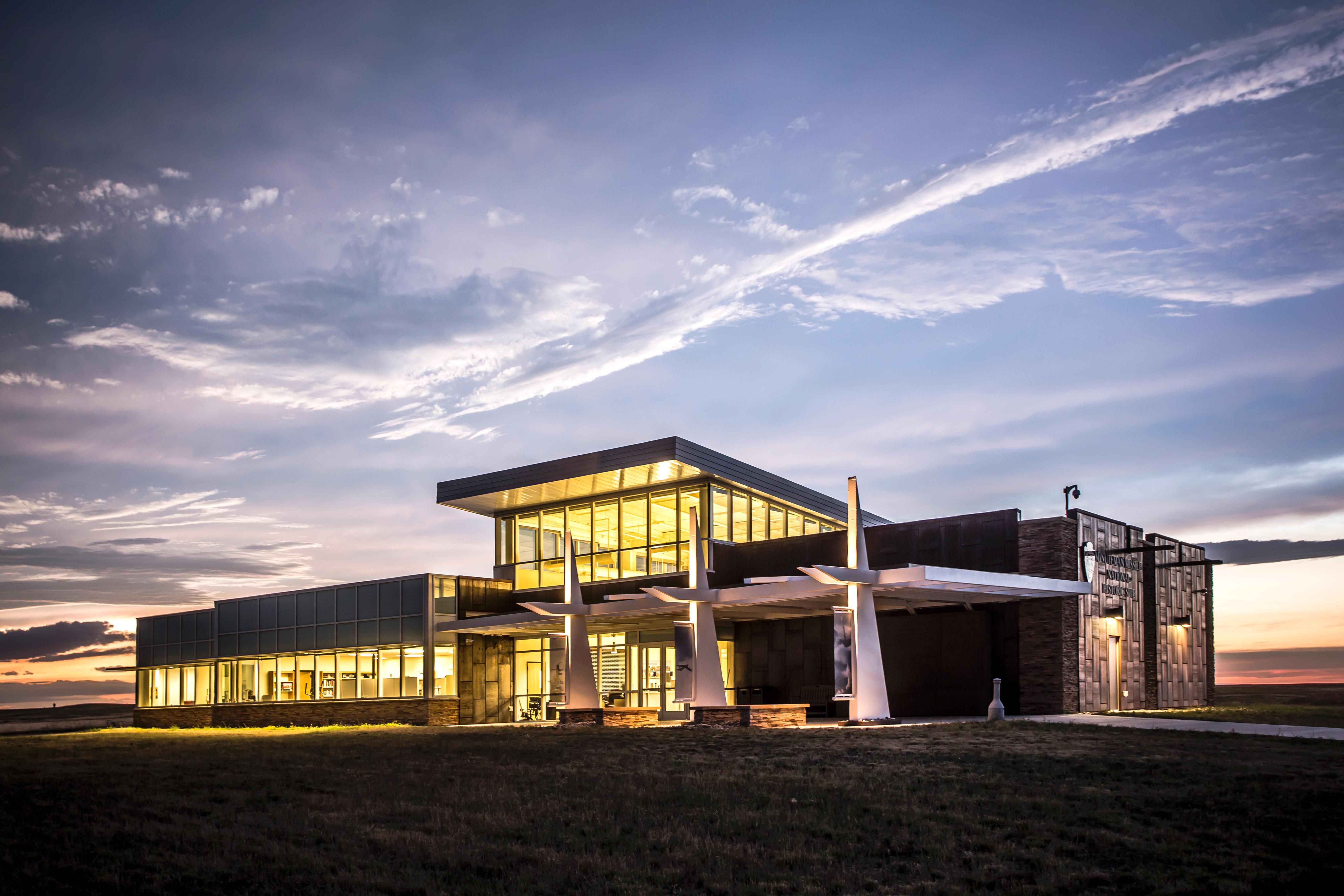 A modern building with a prominent glass clerestory dominates a prairie landscape at sunset