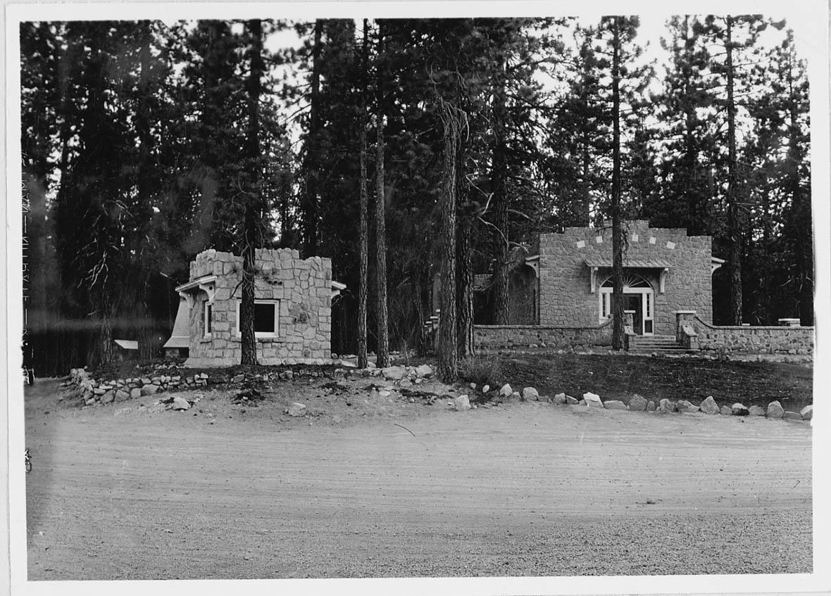 Two one-story stone buildings amid conifer trees fronted by a dirt road.