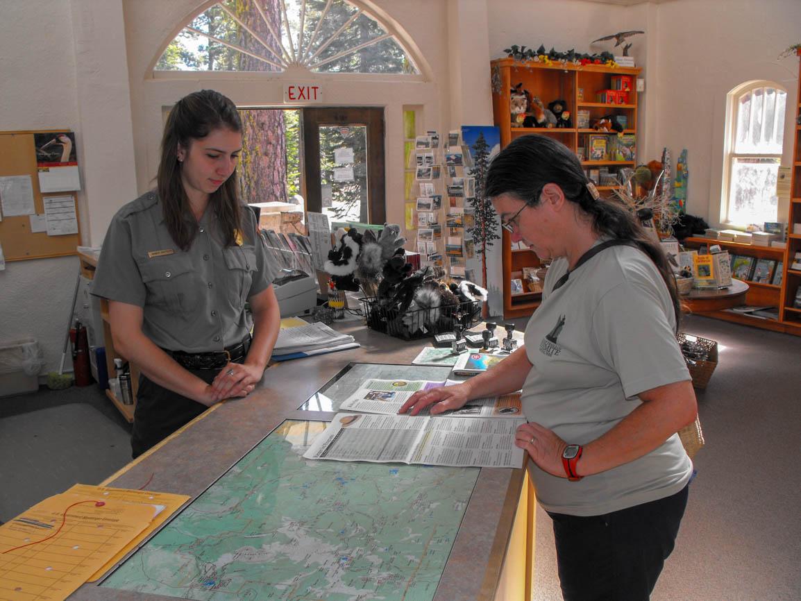 A young woman in a gray uniform shirt and a female visitor stand at a counter inside a museum.