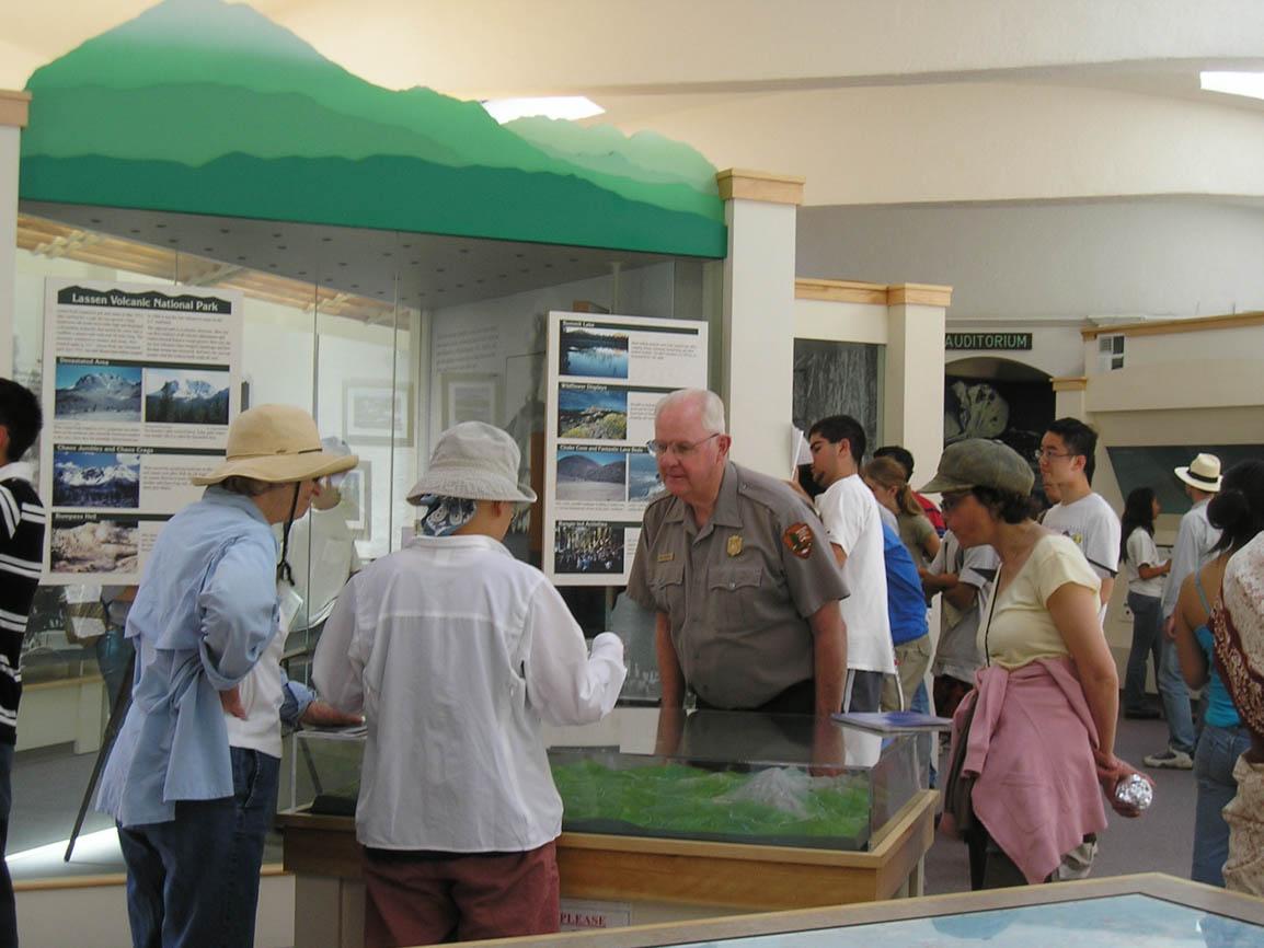 An exhibit hall filled with people. A male ranger talks with visitors at a 3D map in the foreground.