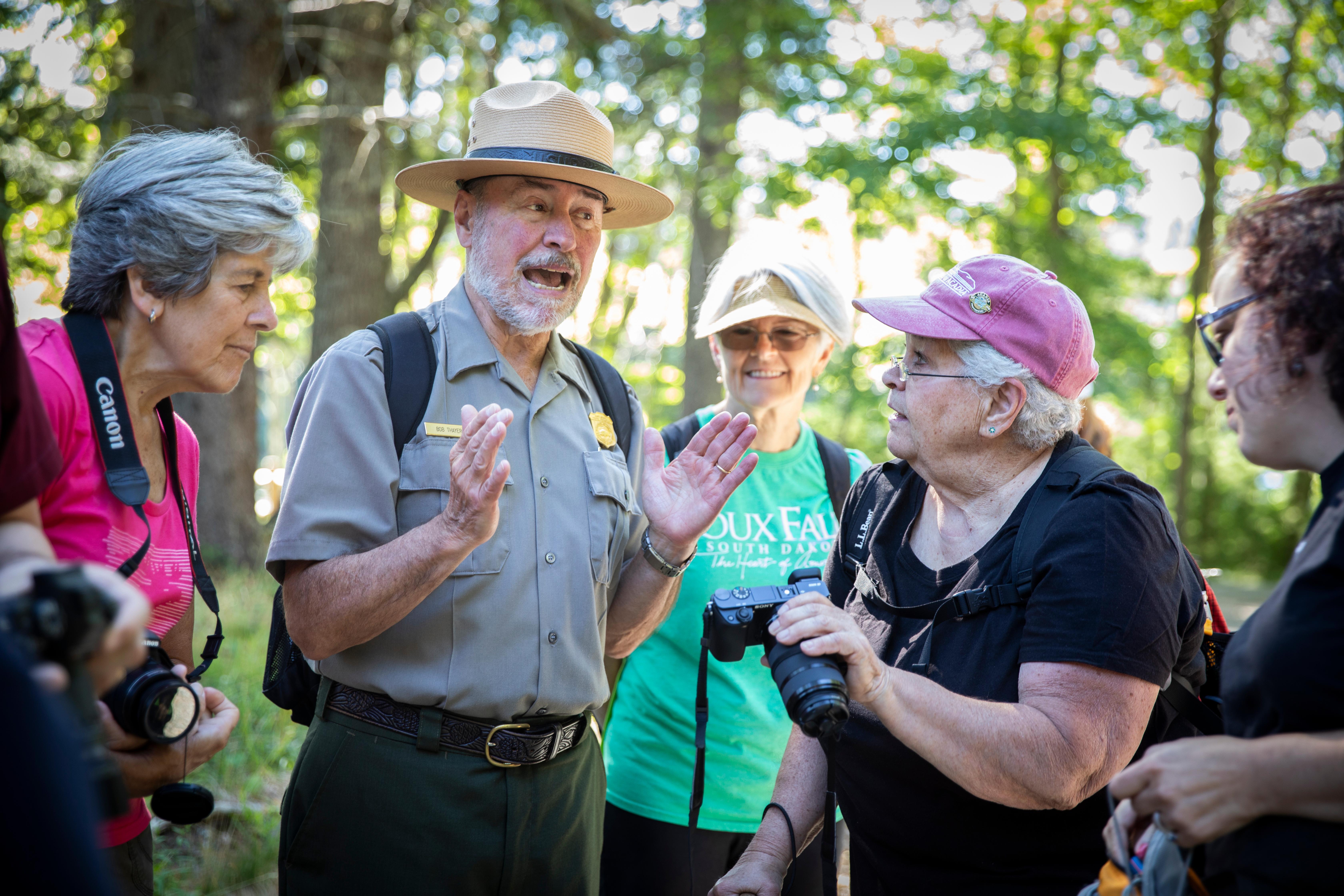 A park ranger in uniform  appears to lead a spirited discussion with a group of visitors in a wooded