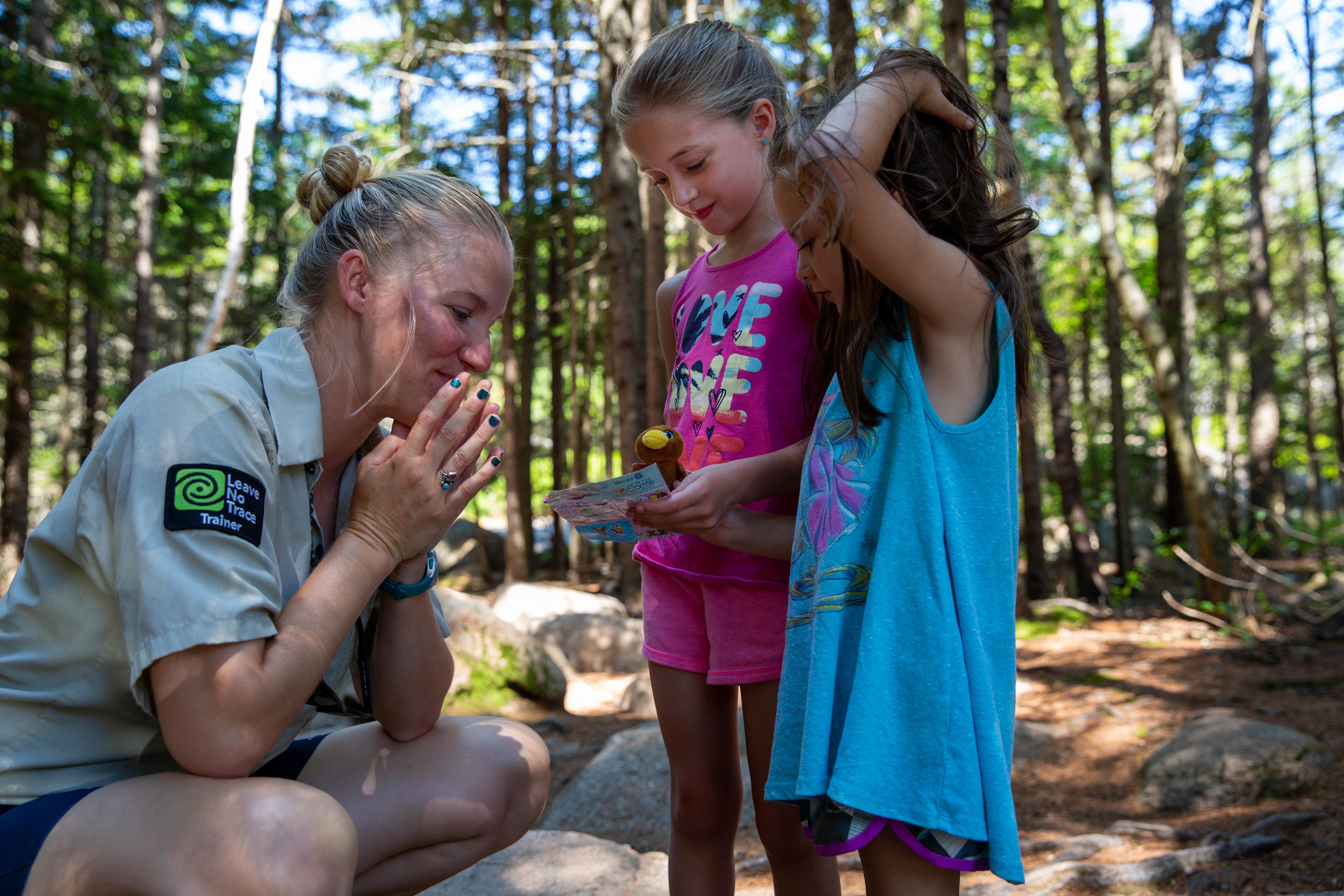 A young woman wearing a uniform shirt crouches low to speak with two young girls holding a small stu