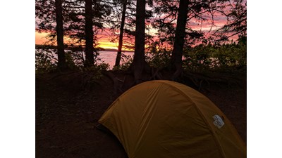 The sun rises through trees on the edge of Feltman Lake. A yellow tent sits in the foreground.