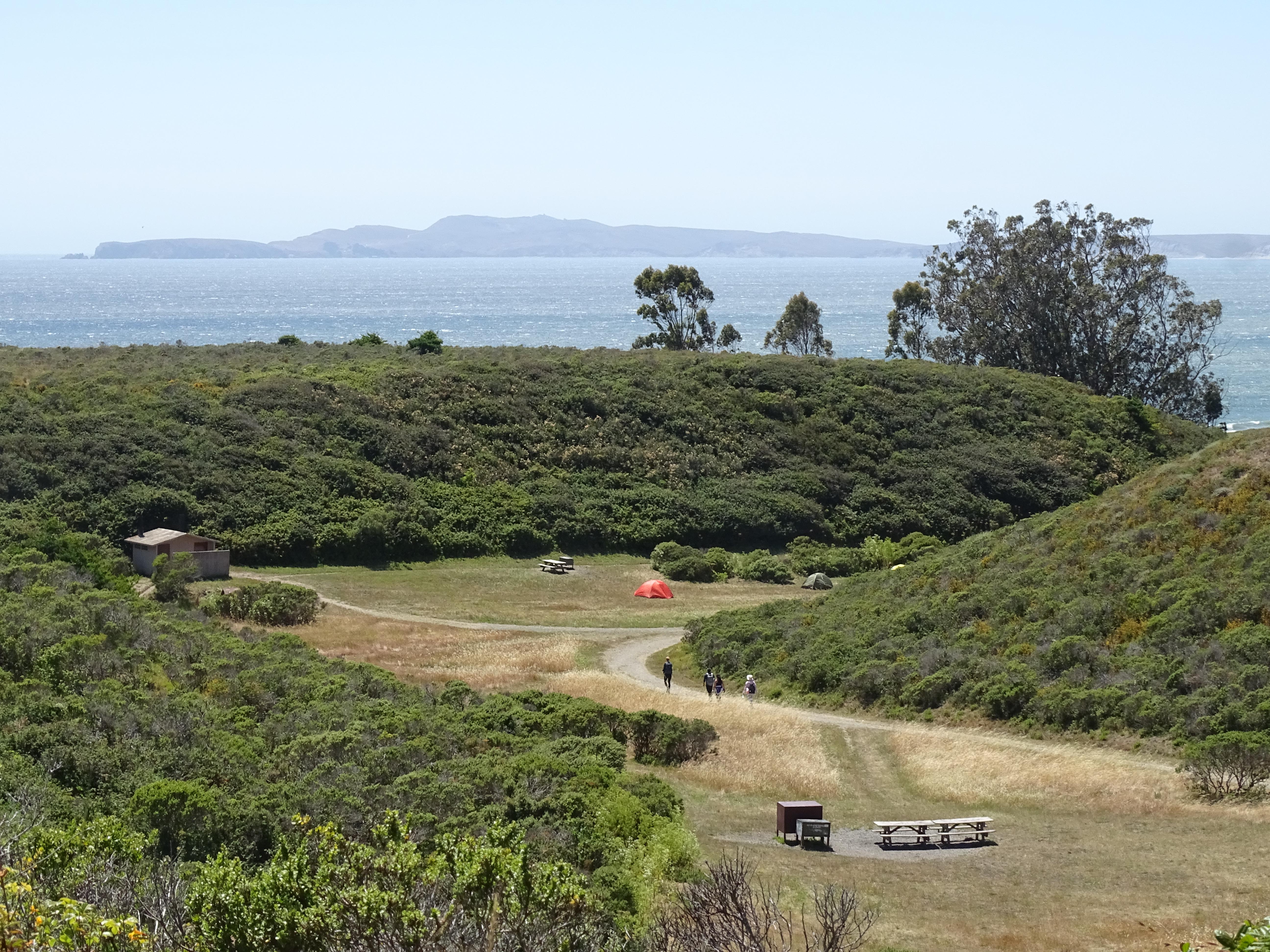 Looking down at a few campsites in a small grassy seaside valley surrounded by shrub-covered hills.