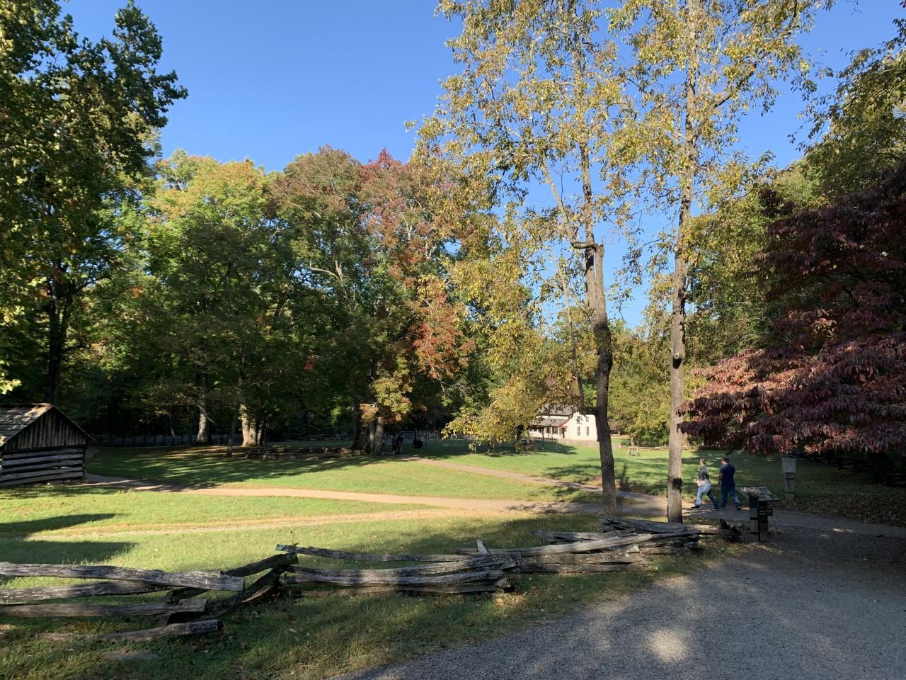 A split rail fence by multiple walkways with two people walking towards buildings and trees.