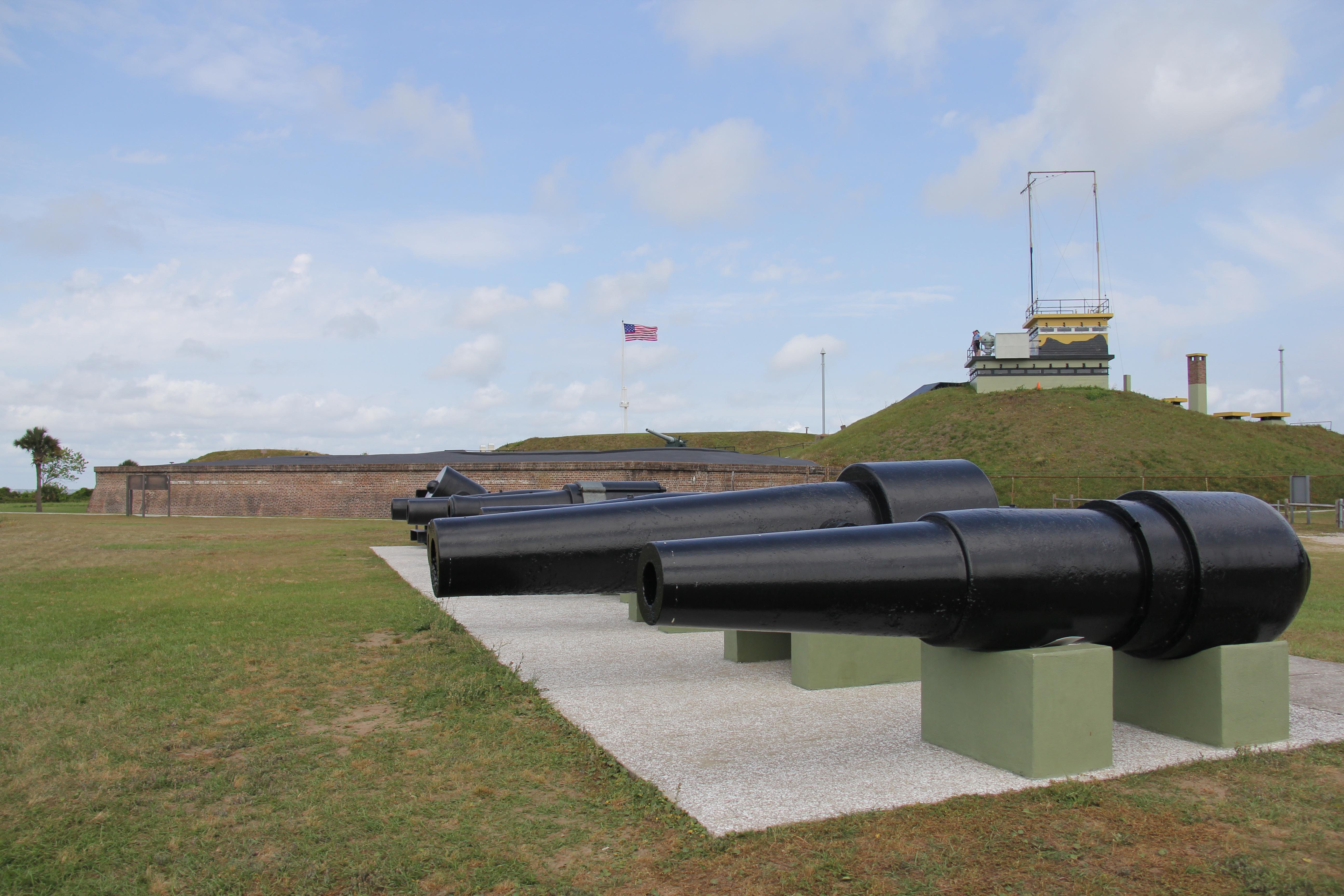 A row of cannon with Fort Moultrie in the background. A US flag is flying in the fort.