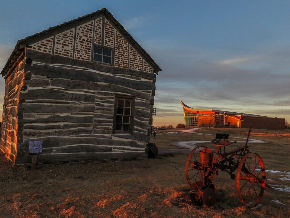 A historic cabin stands next to a concrete path that leads to a plow shaped visitor center