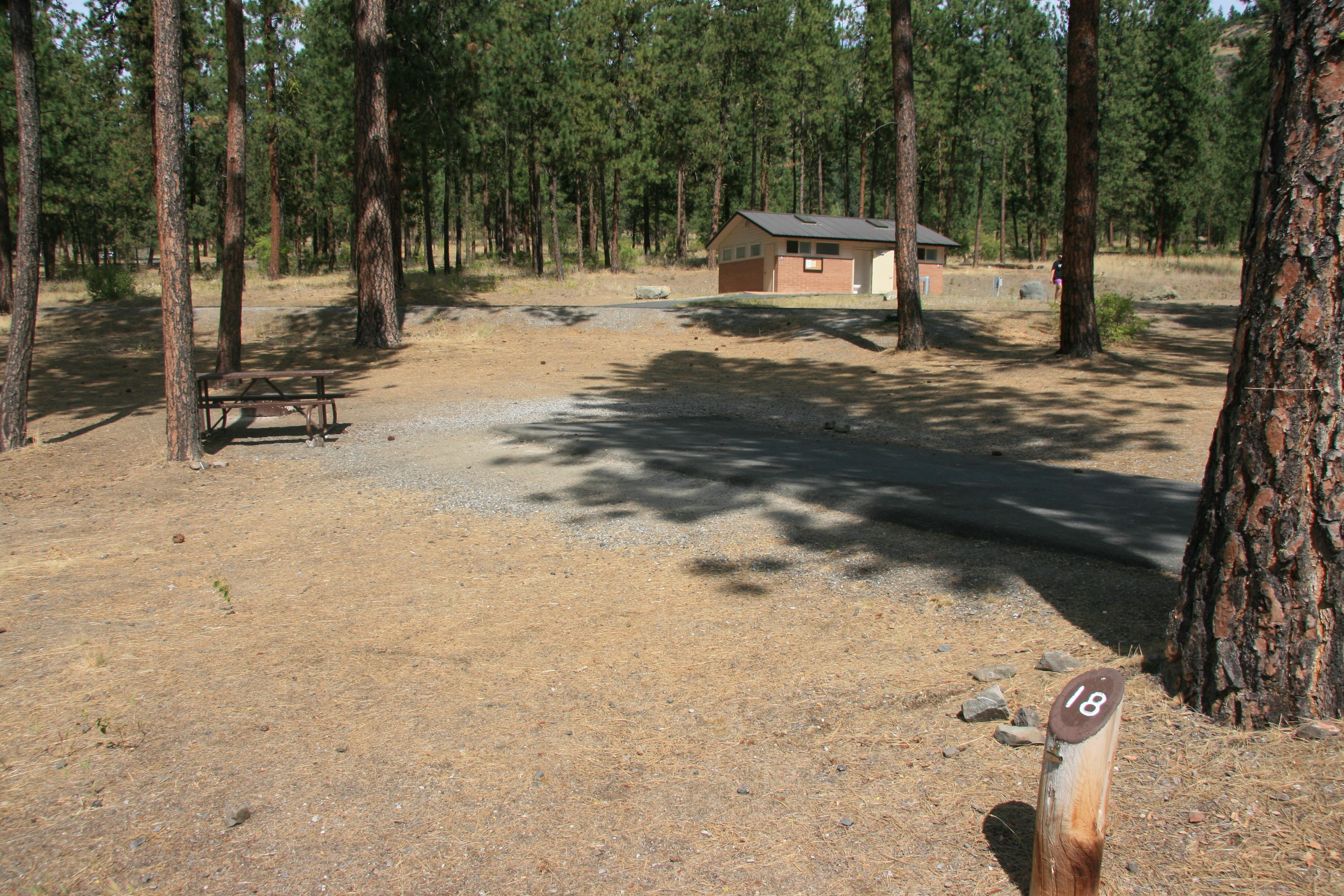 A paved campsite pad with gravel adjacent, a picnic table, and the bathroom building beyond.