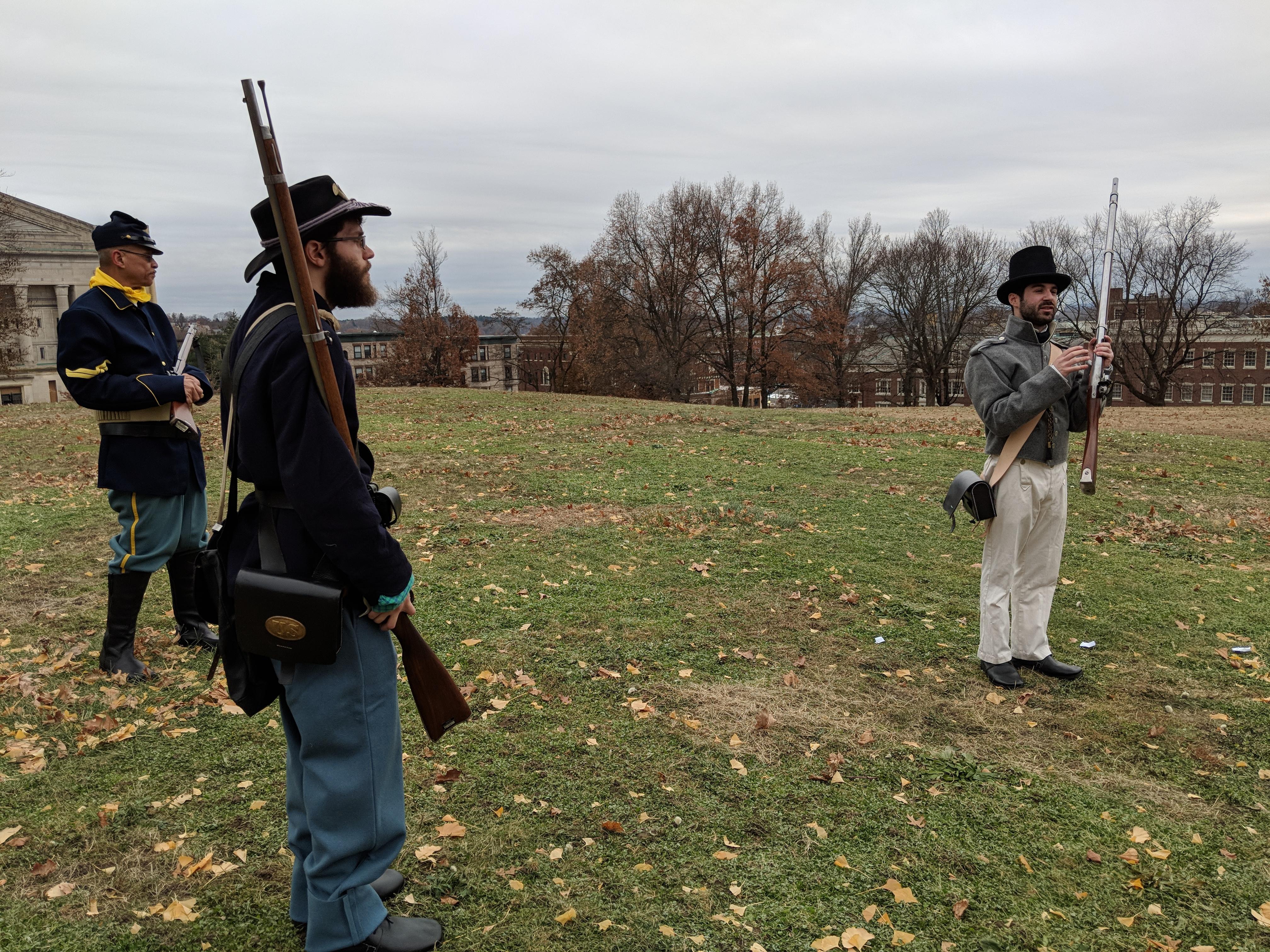 Three Park Volunteers, holding historic firearms, on the grounds of the Armory against a cloudy sky.