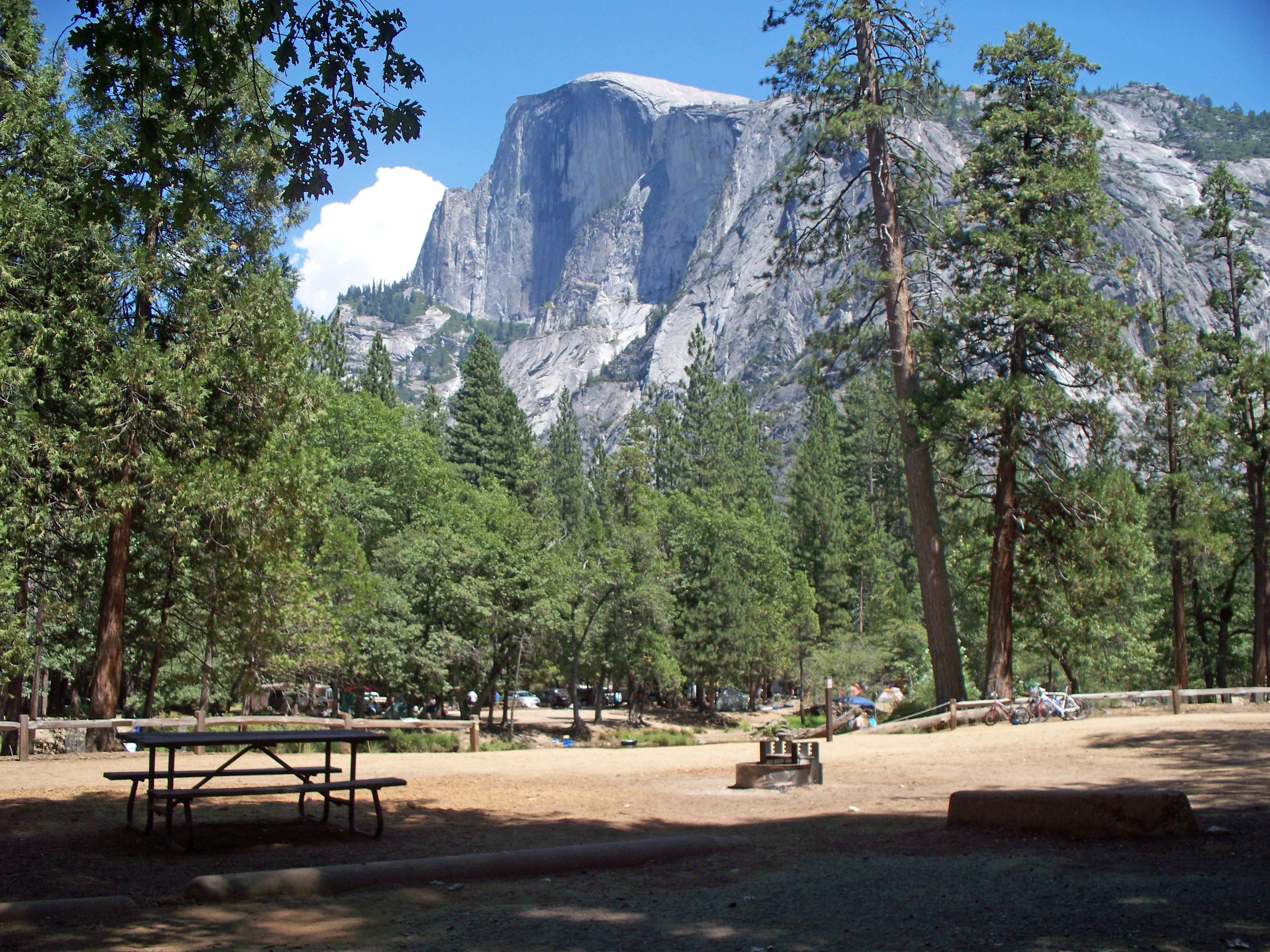 A cleared campsite shows a picnic table and fire pit. A view of Half Dome can be seen through trees.