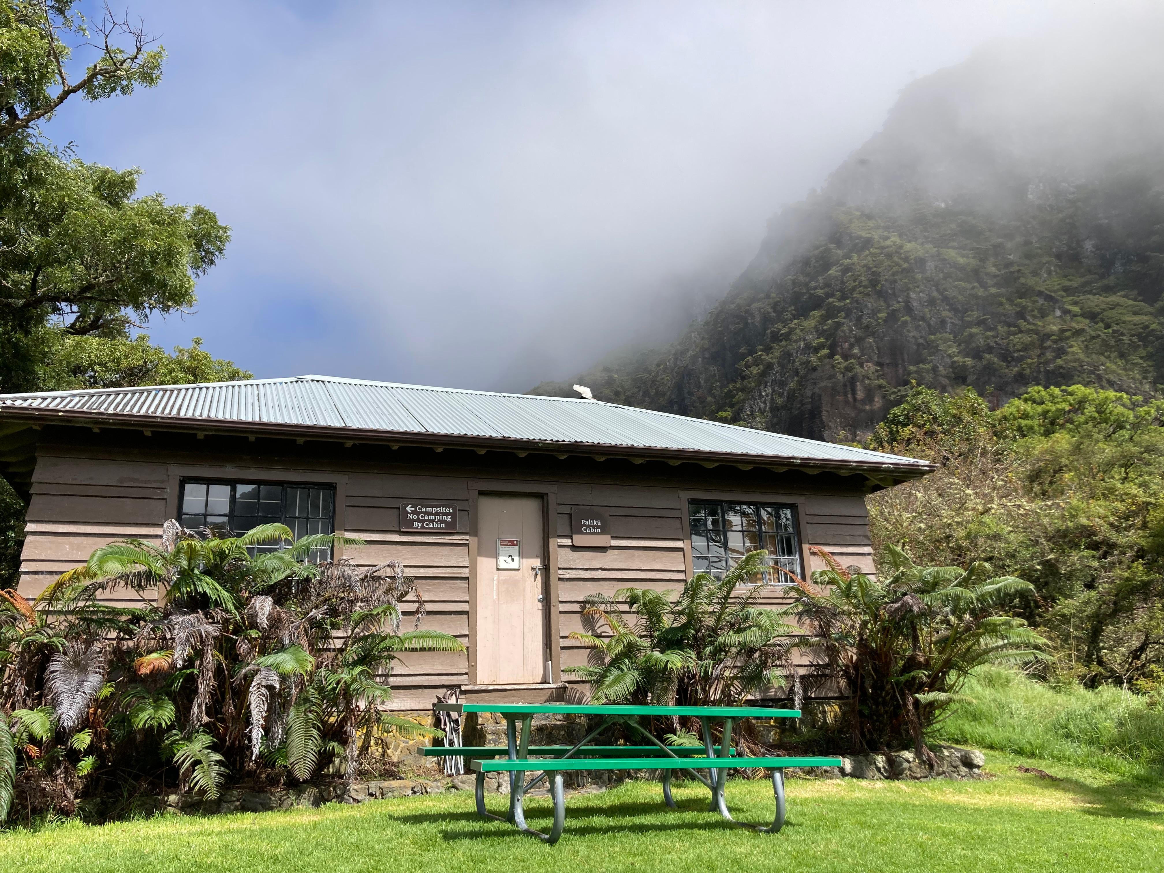 a rustic building with tall cliff and greenery surrounding picnic table in foreground