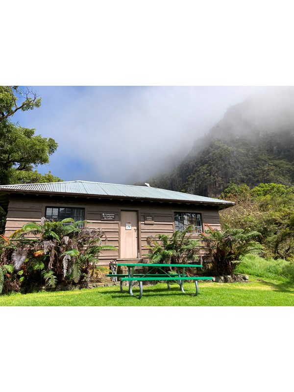 a rustic building with tall cliff and greenery surrounding picnic table in foreground