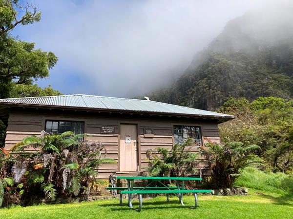 a rustic building with tall cliff and greenery surrounding picnic table in foreground