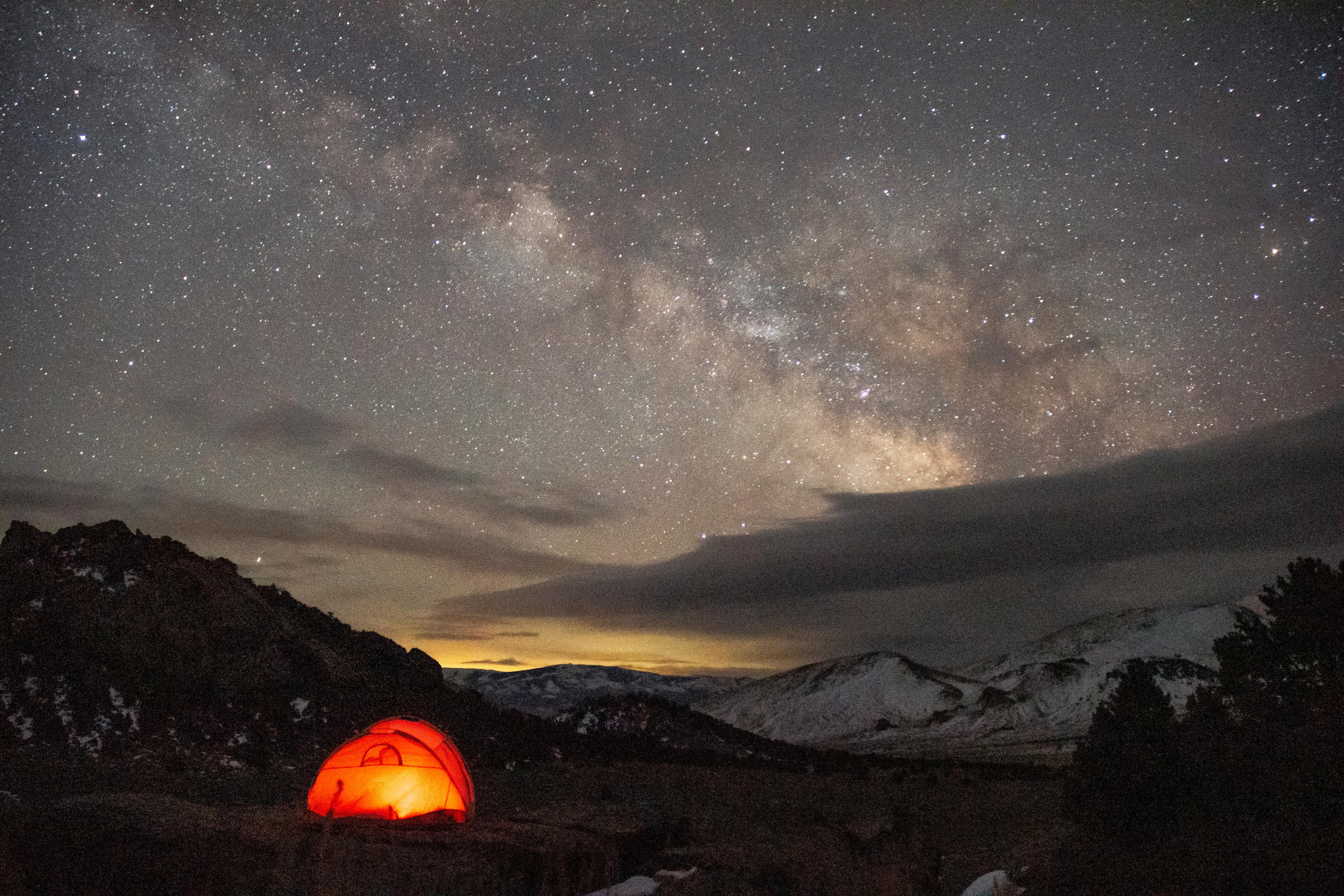 An orange tent glows in early pre-dawn beneath the milky way in a star filled sky.