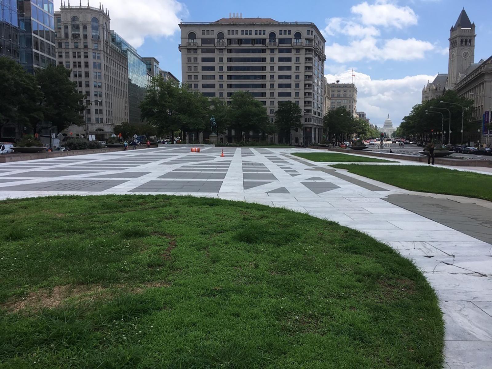 A grass circle in a gray stone plaza with lines resembling streets with the US Capitol in the rear.