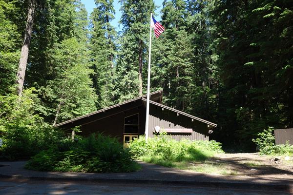 A brown building with a pitched roof sits surrounded by tall old growth trees.