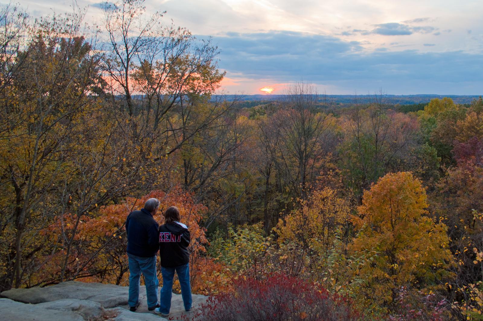 Two people stand at the edge of a gray rock cliff; in the distance, sunset and trees in fall colors.