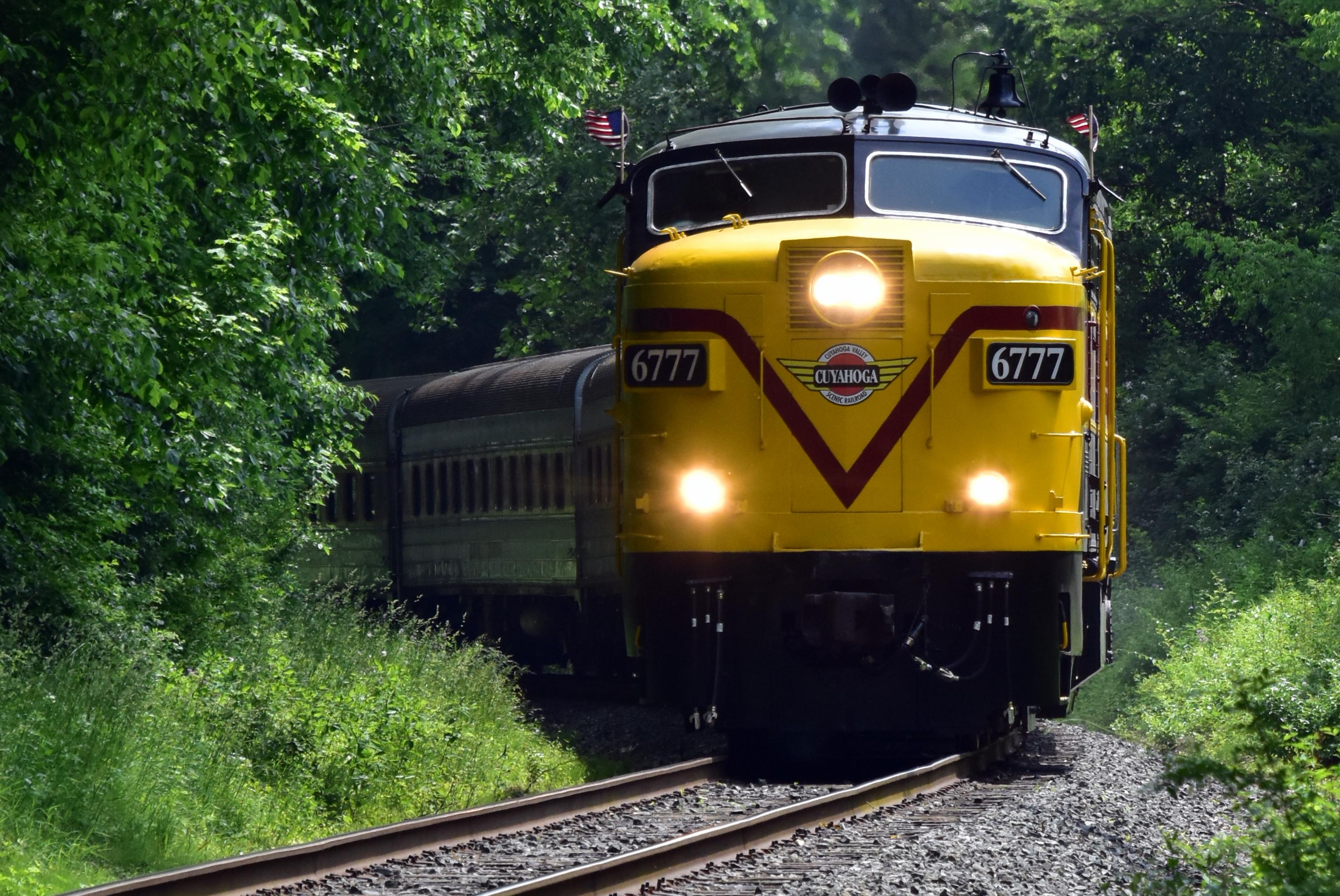 A yellow-and-red engine with number 6777 leads a passenger train along tracks through green trees.