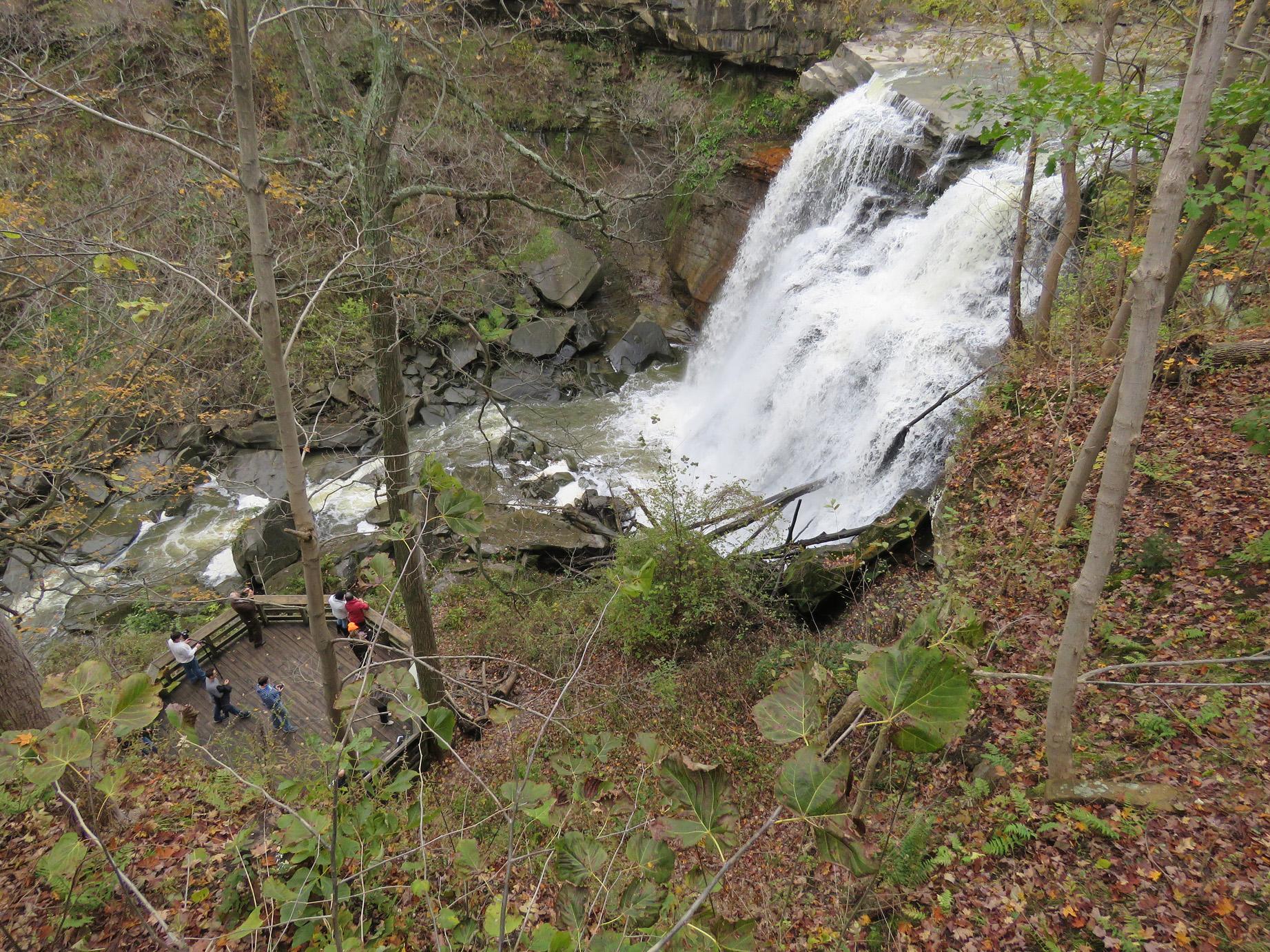 Viewed from above, people stand on a platform near frothy white water falling over a rocky ledge.