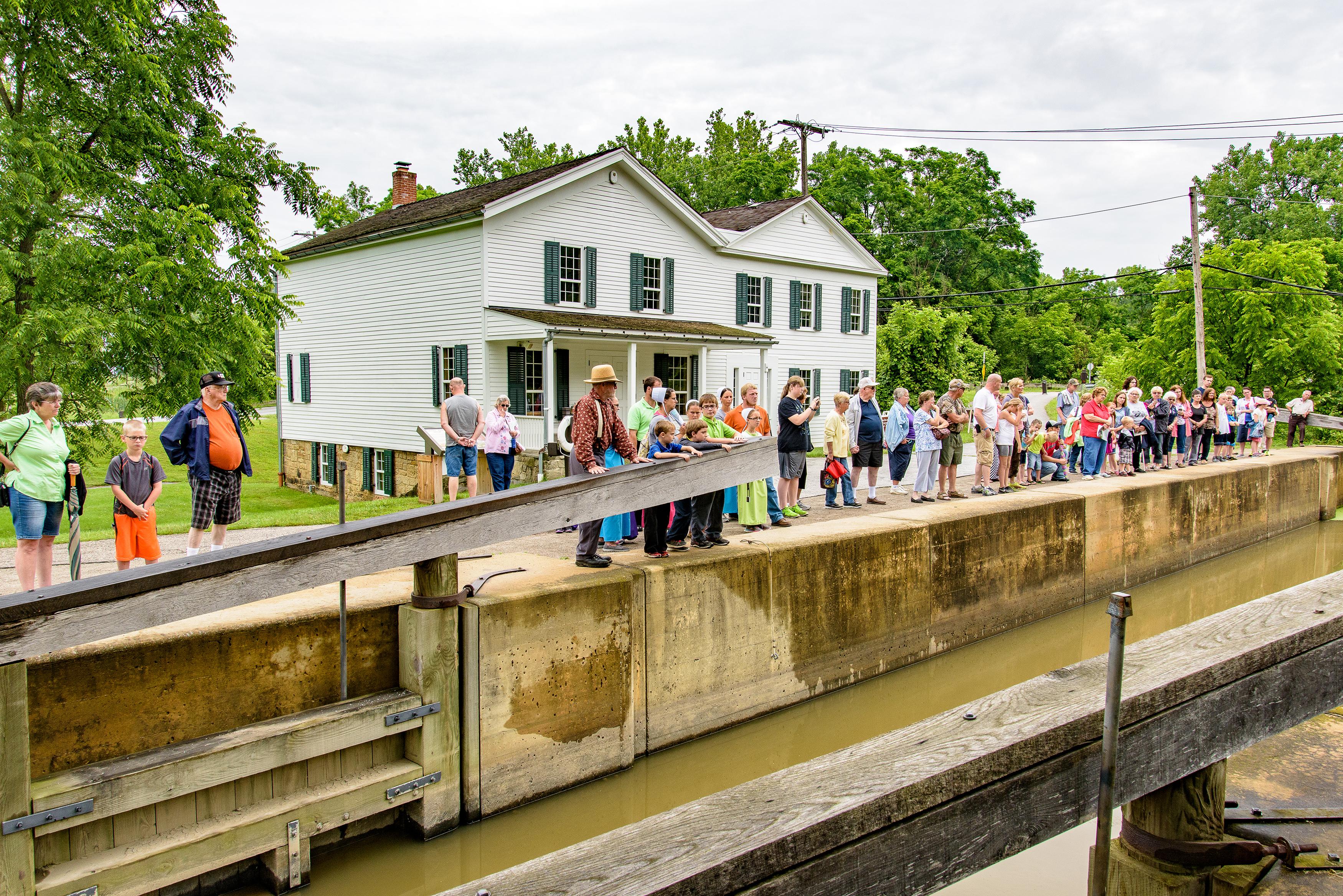 Visitors line the concrete edge of a canal lock filled with water, in front of a white building.