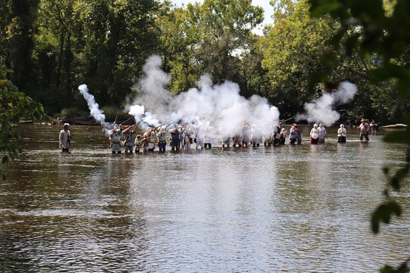 Sycamore Shoals in Elizabethton, TN A large group of living historians stand in a river and fire muskets, with a cloud of smoke rising.