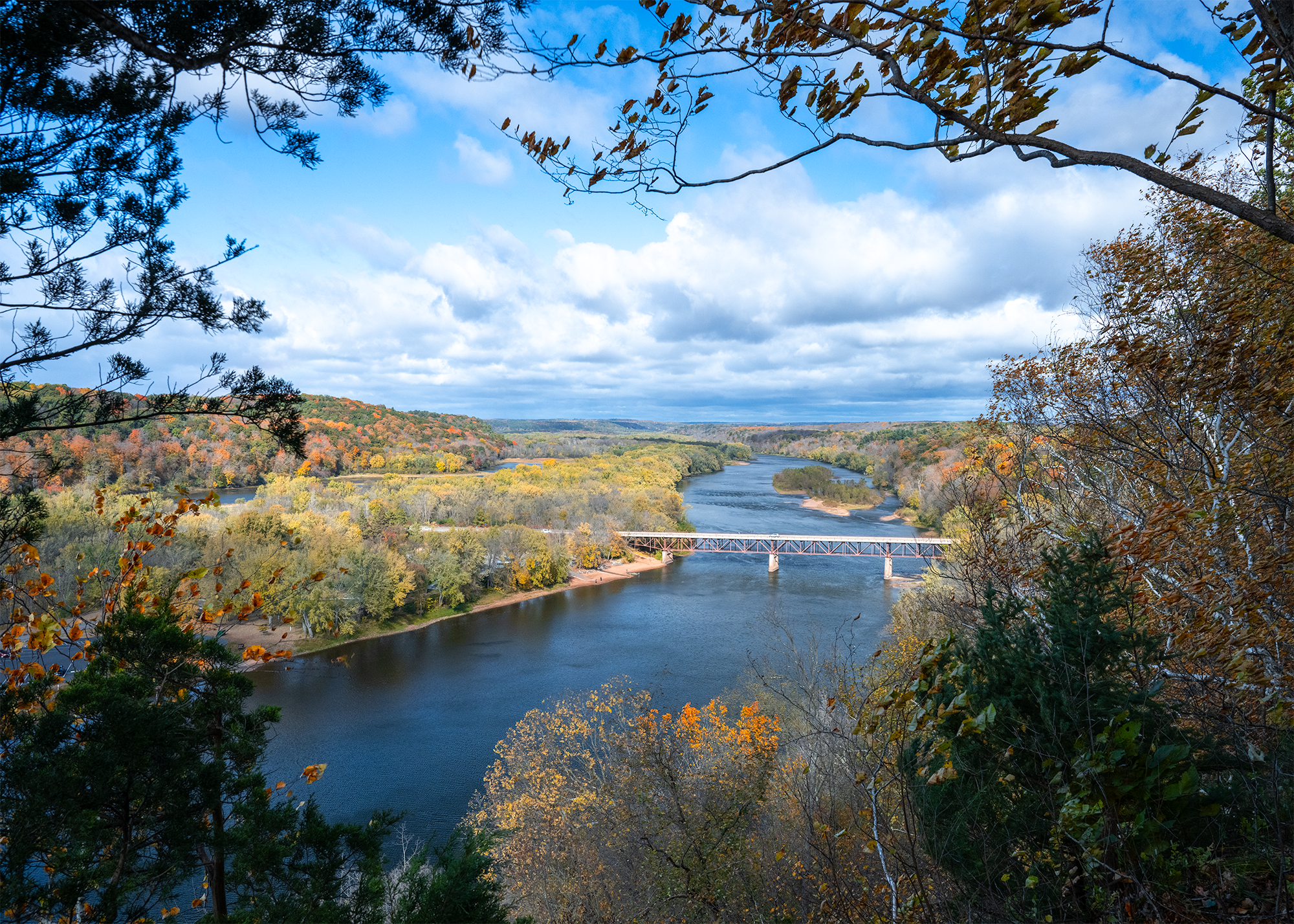 A blue river winds through a valley lined with trees in autumn color.