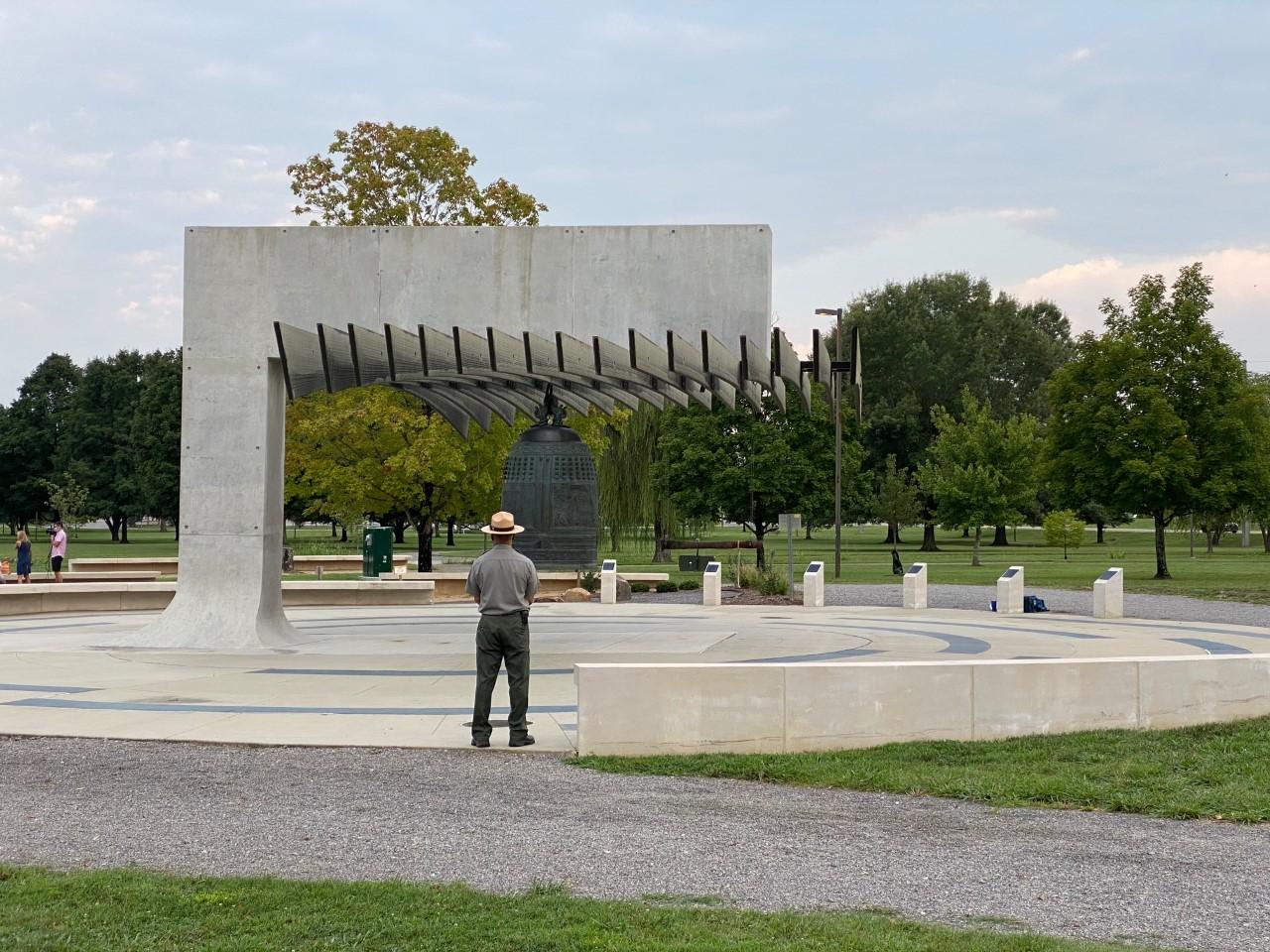 Ranger facing a large bronze cast bell hanging from an abstract pavilion surrounded by green space