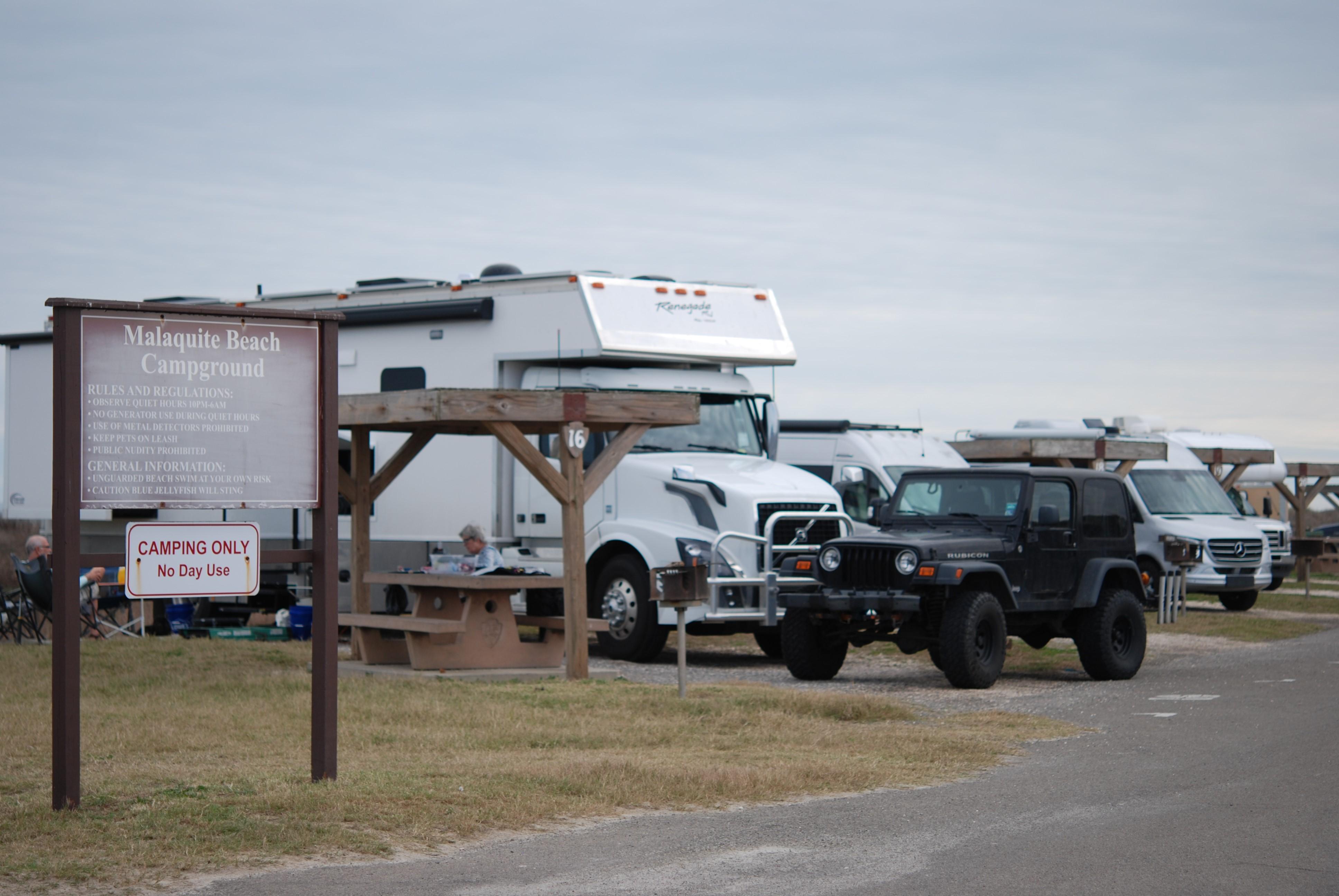 Malaquite Beach Campground sign, camping vehicles in gravel campsites, shade covered picnic table.