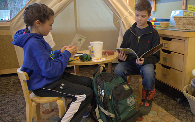 two children sitting at a small table holding books, a green backpack on the floor nearby