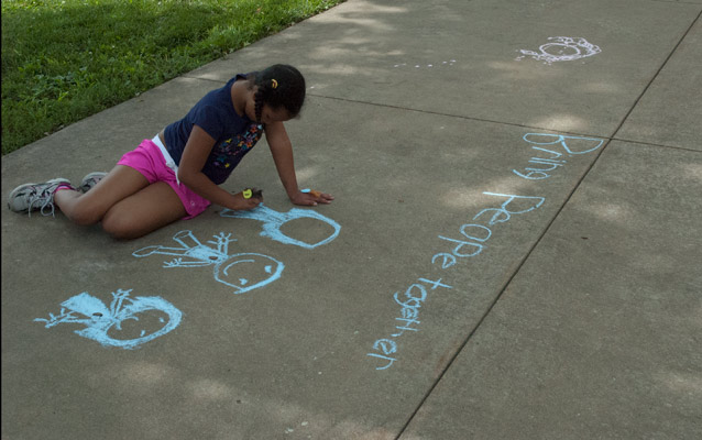 A girl draws a picture of children with sidewalk chalk that says