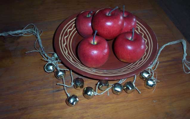 A string of jingle bells and a plate of red apples decorate a wooden table.