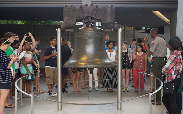 Color photo of teenage visitors gathered around the Liberty Bell in a semi-circle with a male park ranger standing off to the side. Color photo of teenage visitors gathered around the Liberty Bell in a semi-circle with a male park ranger standing off to the side.