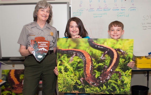 Two kids hold up a large picture of a Shenandoah Salamander next to a park ranger who holds up an arrowhead, the symbol of the National Park Service. Two kids hold up a large picture of a Shenandoah Salamander next to a park ranger who holds up an arrowhead, the symbol of the National Park Service.