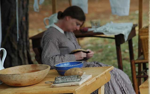 Woman in long dress and hair pulled back knits on a porch next to a wooden table.