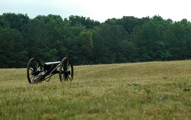 A Civil War era cannon sits in a green field pointing towards a large group of trees.