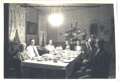 A group of boarders seated around a table eat dinner at a boarding house.