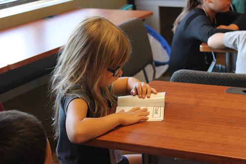 A blond little girl with glasses examines a booklet closely that she is resting on a table