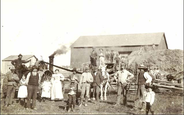 B&W image of farmers, women and children (homesteaders) standing in front of haystack, barn, and smoking engine.  