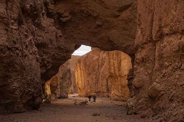 Two people hike below a natural bridge carved out of rock.