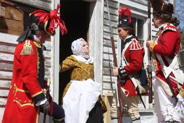Revolutionary War-era British soldiers in red coats confronting an elderly woman at the door of her house.