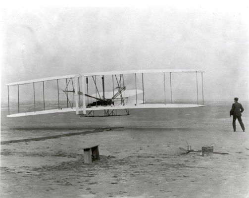 photo of a sandy landscape. A man is at the far right. Center left is a man laying flat on a bi-wing plane that is lifting up off the ground 