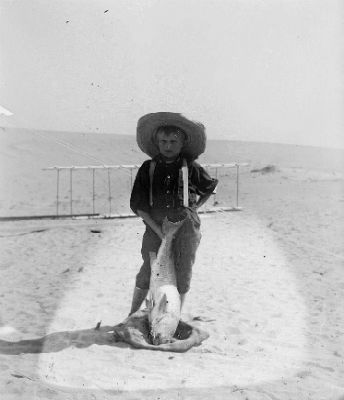 photo of a young boy on a beach in front of a box kite. He has on a sun hat and his right hand on his hip. In his left he is holding a fish.