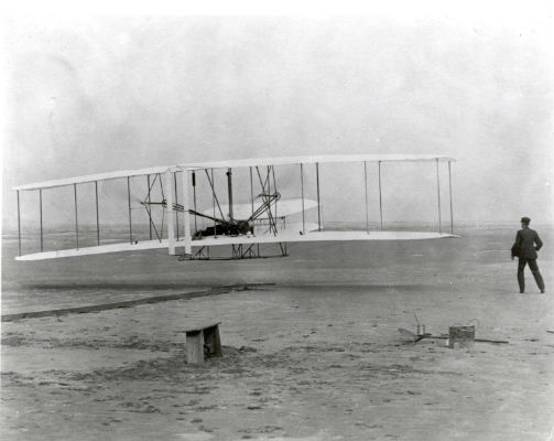 photo of a sandy landscape, to the left a flying machine is lifting off the ground, a man laying in the center. To the right another man stands. photo of a sandy landscape, to the left a flying machine is lifting off the ground, a man laying in the center. To the right another man stands.