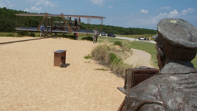 color photo of a bronze sculpture of the wright flyer on the left and in the right corner a statue of a man with a camera