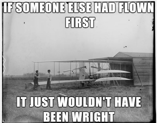 black and white photo of a flying machine next to a wooden hanger with two men standing by the left wing