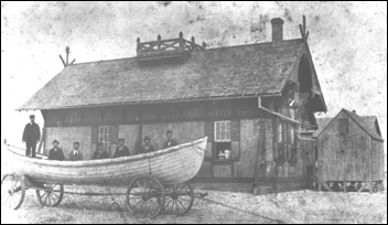 A wooden rowboat sits on wheels in front of a four-wall, hipped roof building. Seven men stand between the boat and the building.