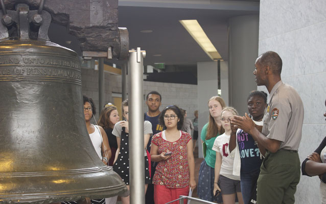 Color photo showing a male park ranger speaking to a group of teens standing in front of the Liberty Bell.