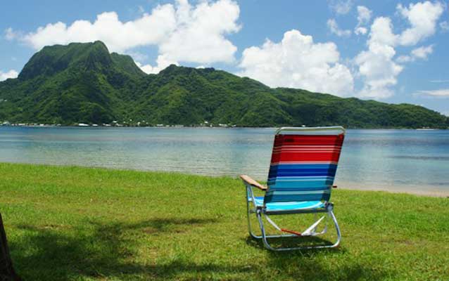 A beach chair sitting along the water's edge with blue skies and green mountains in the distance A beach chair sitting along the water's edge with blue skies and green mountains in the distance
